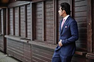 Elegant indian macho man model on suit and pink tie posed against wooden stalls. photo