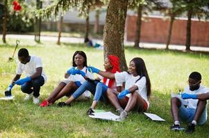 grupo de felices voluntarios africanos sentados bajo un árbol en el parque y escribiendo algo en portapapeles. Concepto de voluntariado, caridad, personas y ecología de África. foto