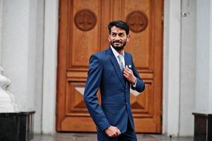 hombre indio de barba elegante con bindi en la frente, vestido con traje azul posado al aire libre contra la puerta del edificio. foto