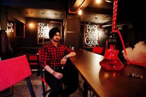 Indian man in checkered shirt and black turban sitting at bar with hookah. photo