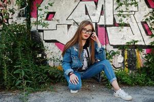 chica hipster casual con estilo en ropa de jeans y gafas contra una gran pared de graffiti. foto