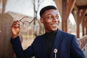 un hombre afroamericano de aspecto asombroso usa un blazer azul con broche, cuello alto negro y anteojos posados en la calle. chico negro de moda. foto