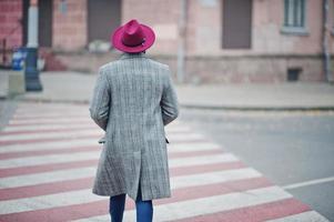 Back of walking stylish African American man model in gray coat and red hat at crosswalk. photo