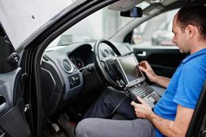 Car repair and maintenance theme. Electric mechanic in uniform working in auto service, making car diagnostics using obd device with laptop. photo