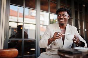 Stylish afro man in beige old school suit sitting on cafe with cup of coffee. Fashionable young African male in casual jacket on bare torso. photo