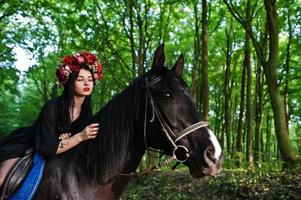 Mystical girl in wreath wear in black at horse in wood. photo