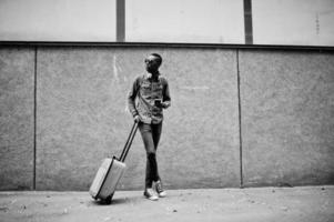 African american man in checkered shirt, sunglasses and earphones with suitcase. Black man traveler against wall holding mobile phone. photo