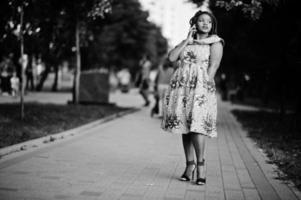 linda chica afroamericana de pequeña altura con rastas, vestida con un vestido amarillo de color, posada al atardecer, caminando y hablando por teléfono celular. en blanco y negro. foto