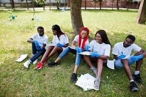 grupo de felices voluntarios africanos sentados bajo un árbol en el parque y escribiendo algo en portapapeles. Concepto de voluntariado, caridad, personas y ecología de África. foto