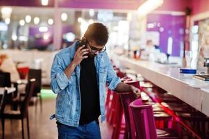 Elegante hombre asiático vestido con chaqueta de jeans y anteojos posados contra el bar en el club y hablando por teléfono móvil. foto