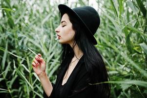 Sensual smoker girl all in black, red lips and hat. Goth dramatic woman smoking on common reed. photo