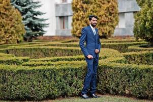 Stylish beard indian man with bindi on forehead, wear on blue suit posed outdoor against green bushes at  park. photo