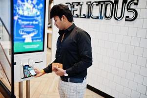 Indian man customer at store place orders and pay by contactless credit card on mobile phone through self pay floor kiosk for fast food, payment terminal. Pay pass. photo