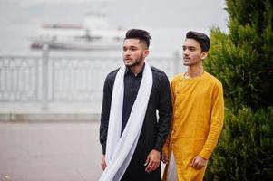 Two indian stylish mans friends in traditional clothes posed outdoor against steamboat on sea. photo
