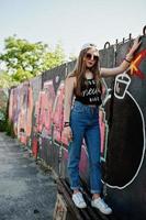 elegante chica hipster casual con gorra, gafas de sol y jeans desgastados contra una gran pared de graffiti con una gran bomba tnt. foto