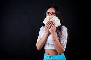 retrato de una chica atractiva con camiseta azul o turquesa, pantalones y gafas posando con mucho dinero en la mano. foto