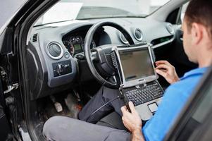 Car repair and maintenance theme. Electric mechanic in uniform working in auto service, making car diagnostics using obd device with laptop. photo