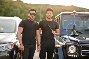 Two asian brothers man wear on all black posed near suv cars and smoke hookah. photo