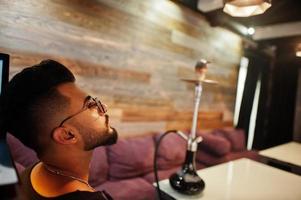 hombre árabe de barba elegante con gafas y camiseta negra fumando narguile en el bar interior. modelo árabe descansando. foto