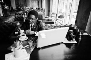 hombre afroamericano de negocios vestido con traje negro y anteojos sentado en la oficina con una computadora portátil y trabajando, vierte azúcar en el café. foto