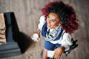 atractiva chica rizada afroamericana con blusa blanca y pantalones cortos azules posó en el café con café con leche y teléfono móvil a mano. vista desde arriba. foto