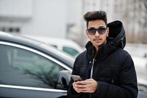 Casual young indian man in black jacket and sunglasses posed against suv car with phone at hands. photo