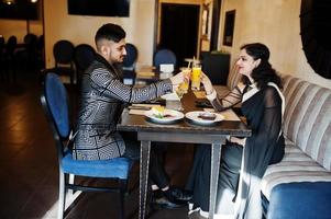 Lovely indian couple in love, wear at saree and elegant suit, sitting on restaurant and cheers together by orange juice. photo