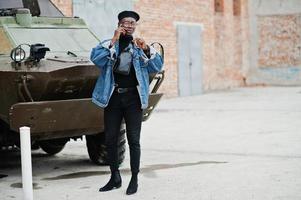 African american man in jeans jacket, beret and eyeglasses, with cigar posed against btr military armored vehicle, and speaking on mobile phone. photo