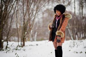 Curly hair african american woman wear on sheepskin coat and gloves posed at winter day throws up snow. photo