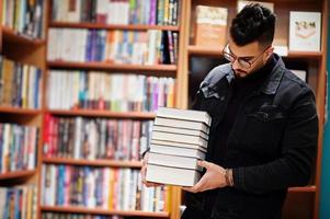 estudiante árabe alto e inteligente, vestido con chaqueta de jeans negros y anteojos, en la biblioteca con una pila de libros. foto