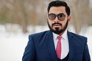 primer plano retrato de elegante hombre de negocios de barba india en traje y gafas de sol posado en el día de invierno al aire libre. foto