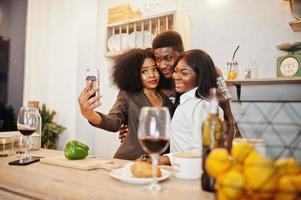 Three african american friends spending time at kitchen with wine. Black peoples relaxing at home and making selfie on phone. photo