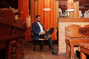 Handsome well-dressed arabian man with glass of whiskey and cigar posed at pub. photo