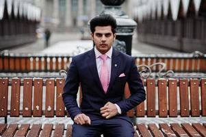 Elegant indian macho man model on suit and pink tie posed on winter day and sitting on bench. photo