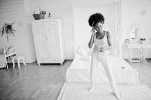 Young african american woman in pink singlet dancing and listen music on earphones at her white room. photo