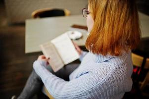 parte trasera de una joven y alegre mujer pelirroja con gafas sentada en su lugar de trabajo en un café leyendo algo en un cuaderno. foto