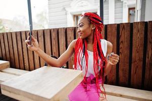 chica afroamericana de moda con pantalones rosas y rastas rojas posadas al aire libre con teléfono móvil. foto