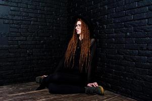Studio shoot of girl in black with dreads, at glasses and hat on brick background. photo