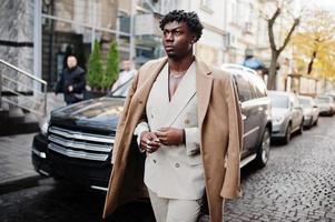 Stylish afro man in beige old school suit and coat walking against black business car. Fashionable young African male in casual jacket on bare torso. photo