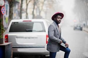 Stylish African American man model in gray jacket tie and red hat posed against silver car suv. photo