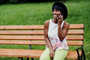 Increíble mujer modelo afroamericana con pantalones verdes y sombrero negro posada en un banco en el parque y hablando por teléfono móvil. foto