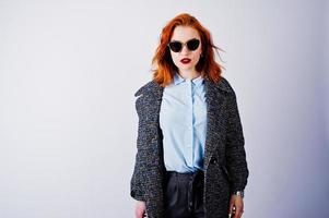 retrato de una fantástica chica pelirroja con camisa azul, abrigo gris posando con gafas de sol en el estudio. foto