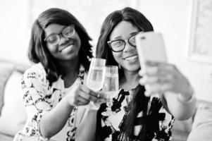 Two african woman friends wear on eyeglasses posed indoor white room, drinking champagne and making selfie. photo