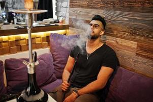 hombre árabe de barba elegante con gafas y camiseta negra fumando narguile en el bar interior. modelo árabe descansando. foto