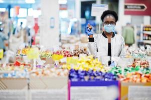 African woman wearing disposable medical mask and gloves shopping in supermarket during coronavirus pandemia outbreak. Black female choose candies at epidemic time. photo