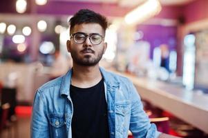 elegante hombre asiático vestido con chaqueta de jeans y gafas posadas contra el bar en el club. foto