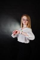 Studio portrait of blonde businesswoman in glasses, white blouse and black skirt against dark background. Successful woman. Girl looking at watches. photo