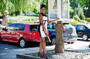 African american girl in sunglasses, black and white dress and handbag posed against cars parking. photo