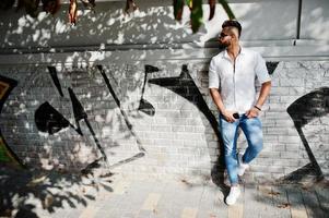 Stylish tall arabian man model in white shirt, jeans and sunglasses posed at street of city. Beard attractive arab guy against graffiti wall. photo