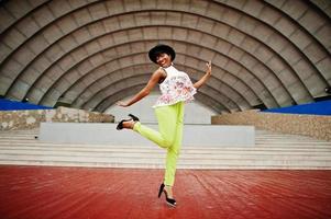 Amazing african american model woman in green pants and black hat posed outdoor against arena hall, jump in the air. photo